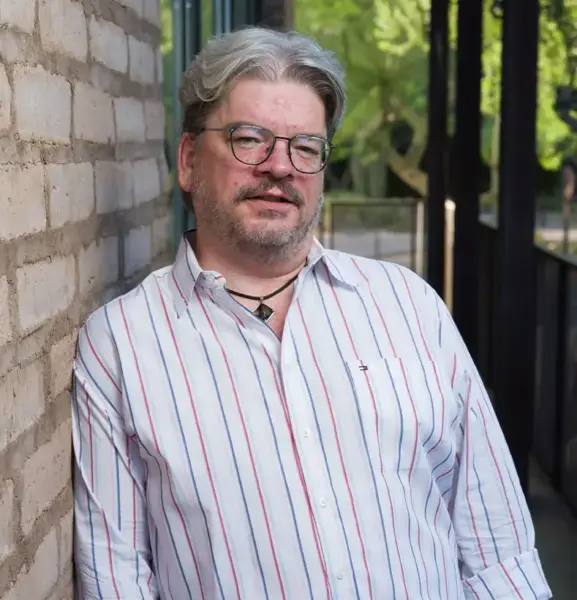 Man with gray hair and glasses leaning against a brick wall outdoors, wearing a white striped shirt and necklace.