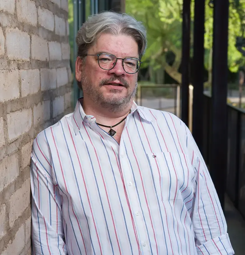 Man with gray hair and glasses leaning against a brick wall outdoors, wearing a white striped shirt and necklace.